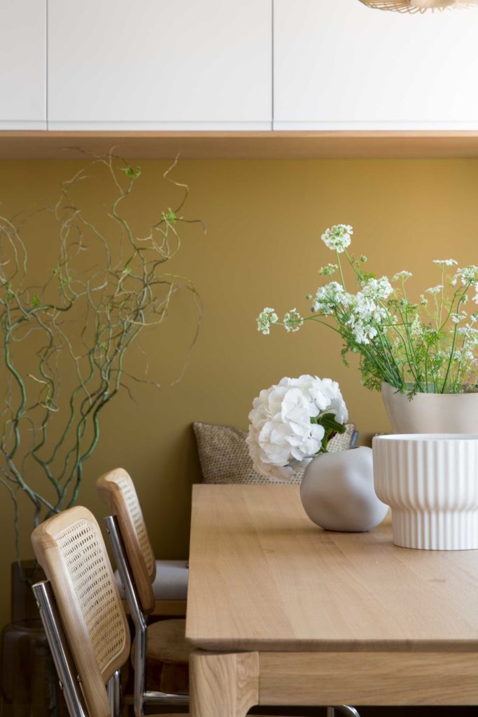 Espace salle à manger de notre projet Jade, table en chêne, chaises en cannage, mur couleur brun cognac. Vases blancs posés sur la table avec des fleurs délicates.