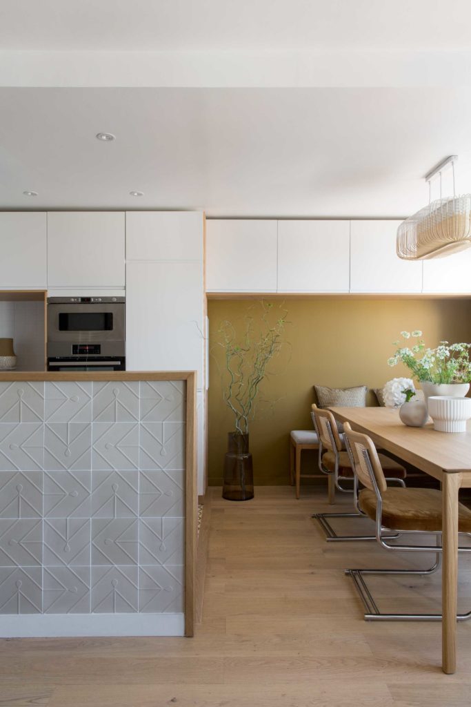 Espace cuisine/salle à manger de notre projet Jade, table en chêne ethnicraft, chaises en cannage NV Gallery, banquette avec coussins et mur du fond peint couleur Brun Cognac. cuisine séparée du salon par un muret habillé de carreau de ciment avec motif beige.