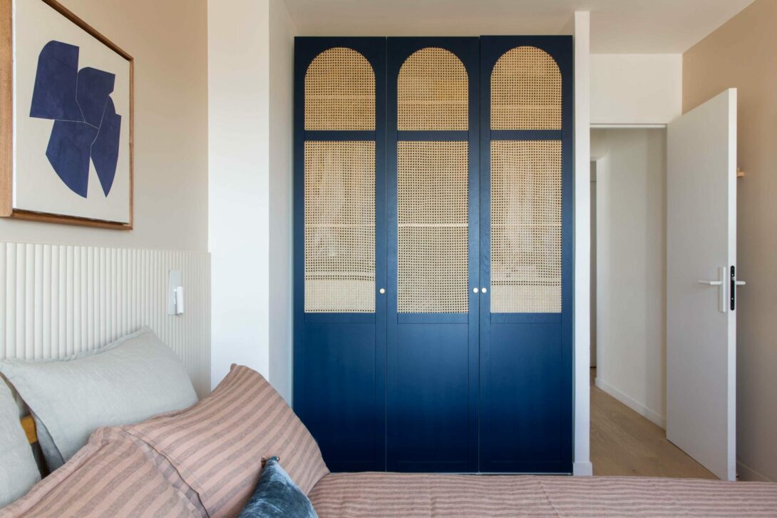 Chambre parentale avec dressing Ikea habillé de portes Plum en cannage et chêne peint bleu. Tête de lit en tasseaux arrondis Orac Décor, liseuse en metal blanc, linge de lit en lin Maison Sauvage, tableau Audrey Noël.