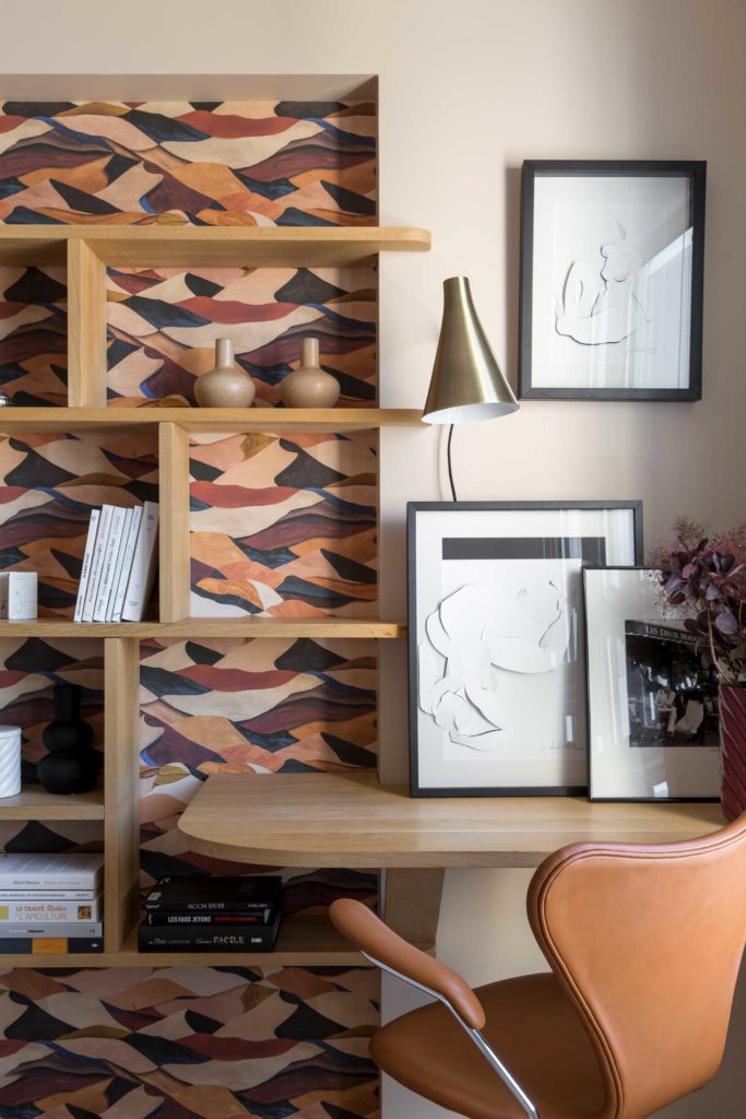 Espace bibliothèque et bureau sur mesure en chêne, papier-peint aux couleur chaudes en fond de bibliothèque, chaise de bureau en cuir Series7 Arne Jacobsen, tableaux posés et suspendus.