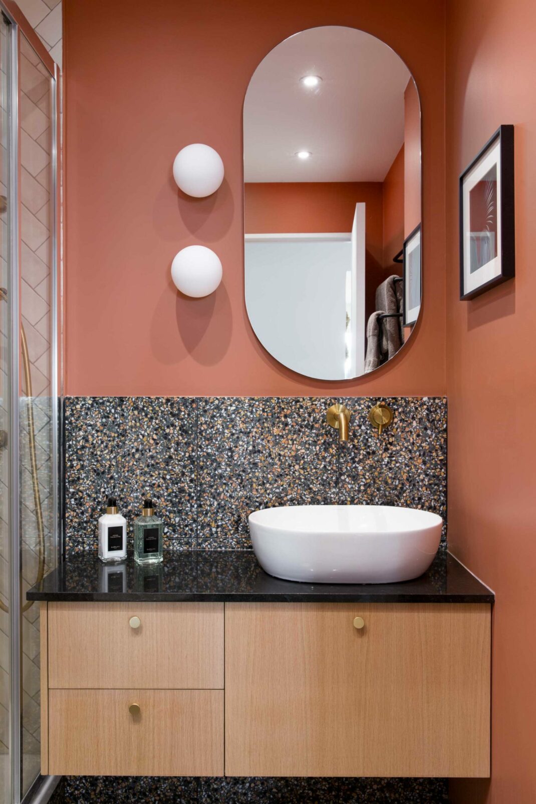 Salle de bain parentale de notre projet Jade, murs peints en terracotta, meuble de salle de bain en chêne, robinet encastré en laiton brossé, plan en granit noir zimbabwé, crédence en terrazzo. Miroir ovale et 2 appliques opalines.