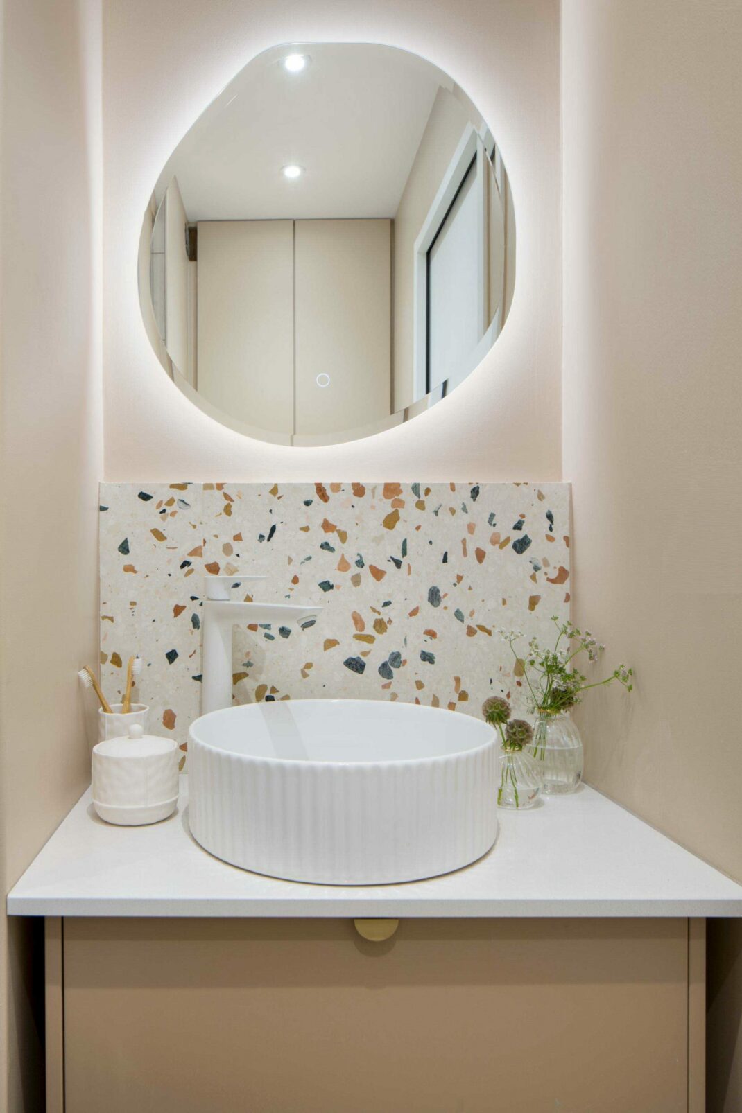 Meuble de salle de bain avec façades Plum couleur Moka, vasque posée en céramique blanche, robinet finition blanc mate, miroir rétroéclairé et crédence en Terrazzo fond beige avec des éclats verts et terracotta.