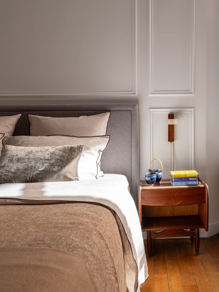 Chambre parentale épurée et chaleureuse, avec une tête de lit capitonnée grise, des coussins en tons neutres et une table de chevet en bois clair. L'applique murale design en bois et travertin et les livres posés sur la table ajoutent une touche de convivialité et de raffinement à cet espace apaisant.