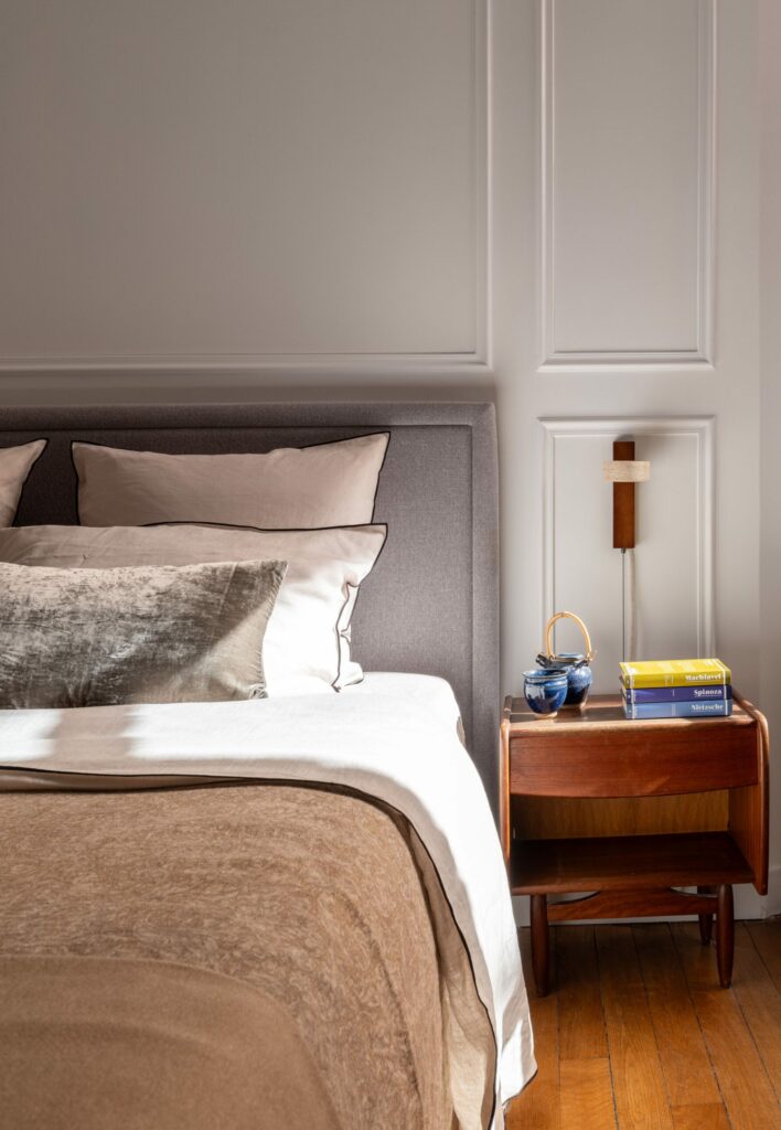 Chambre parentale épurée et chaleureuse, avec une tête de lit capitonnée grise, des coussins en tons neutres et une table de chevet en bois clair. L'applique murale design en bois et travertin et les livres posés sur la table ajoutent une touche de convivialité et de raffinement à cet espace apaisant.