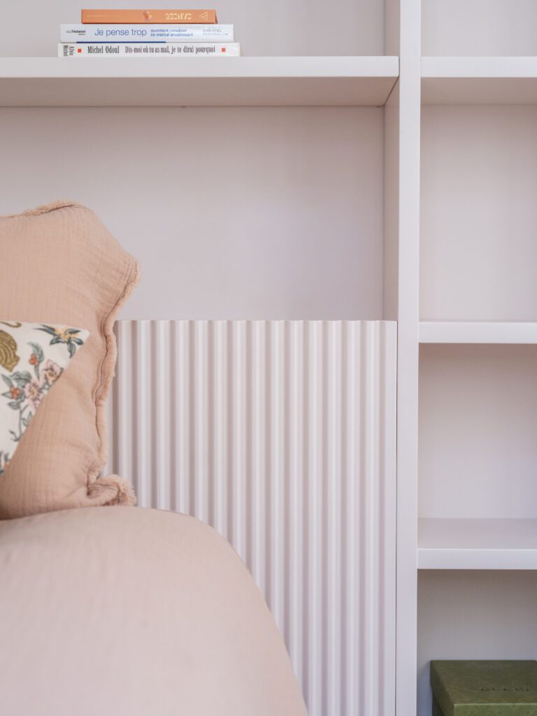 Chambre d'adolescente aménagée avec soin dans un appartement parisien rénové, mettant en valeur un lit confortable avec couverture rose poudré et coussins décoratifs. Rangements sur mesure couleur rose arum avec étagères intégrées, miroir organique et luminaire suspendu élégant. Décoration intérieure moderne et fonctionnelle, alliant couleurs douces, rangements sur mesure optimisés et accessoires décoratifs pour un espace à la fois esthétique et ergonomique.