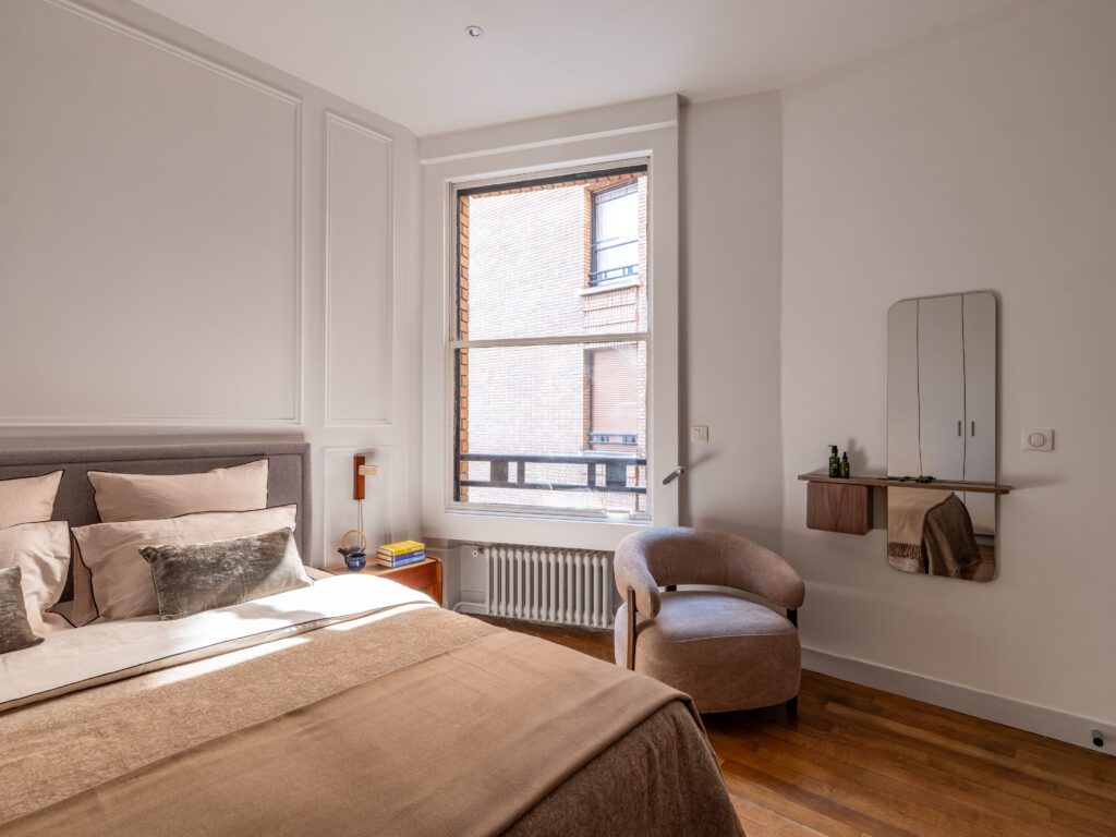 Chambre élégante et lumineuse dans un appartement parisien rénové, avec une tête de lit tapissée gris clair, des coussins assortis et un fauteuil design taupe. Décoration intérieure moderne et chaleureuse, mettant en valeur un miroir mural, une étagette en bois avec produits de soin, et une large fenêtre laissant entrer la lumière naturelle. Sol en parquet et murs blancs pour une ambiance épurée et raffinée.