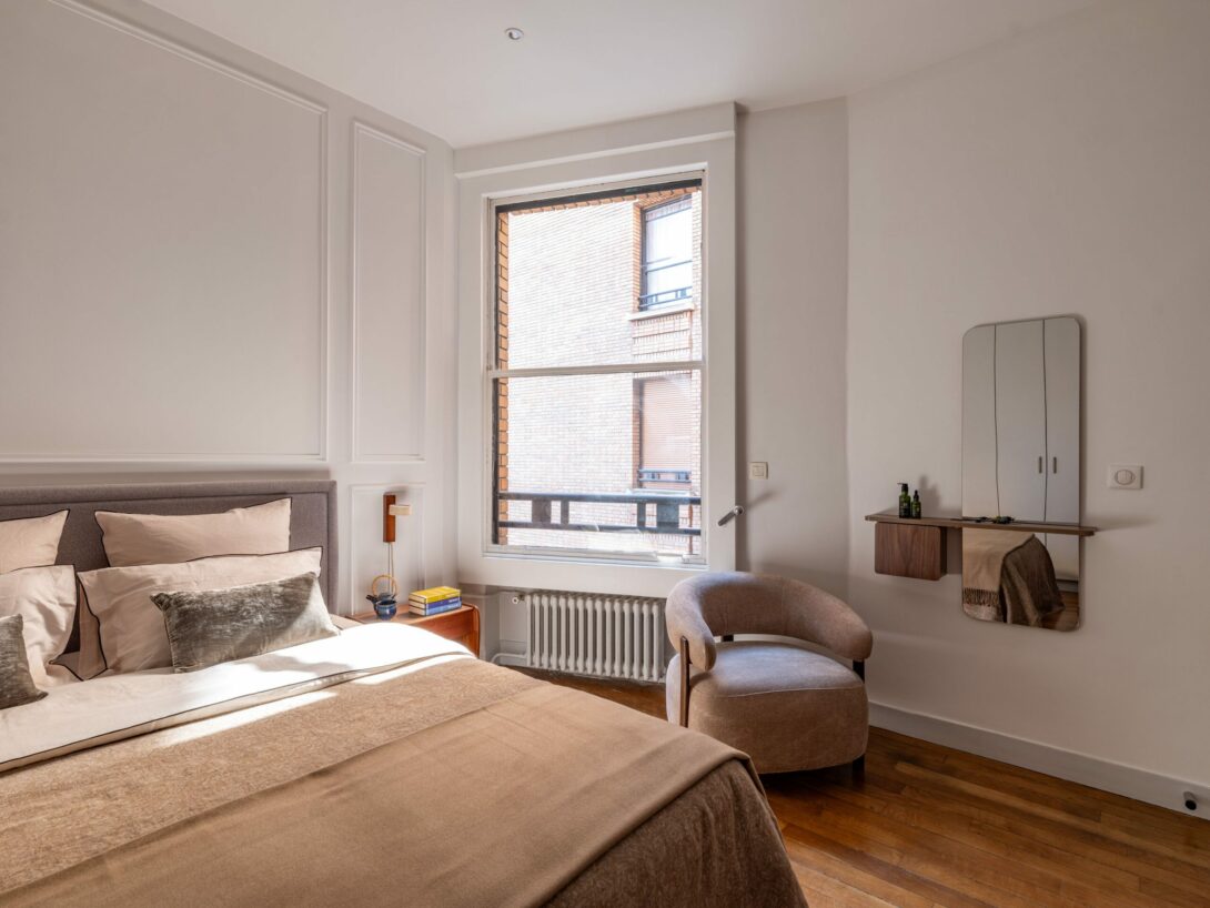 Chambre élégante et lumineuse dans un appartement parisien rénové, avec une tête de lit tapissée gris clair, des coussins assortis et un fauteuil design taupe. Décoration intérieure moderne et chaleureuse, mettant en valeur un miroir mural, une étagette en bois avec produits de soin, et une large fenêtre laissant entrer la lumière naturelle. Sol en parquet et murs blancs pour une ambiance épurée et raffinée.