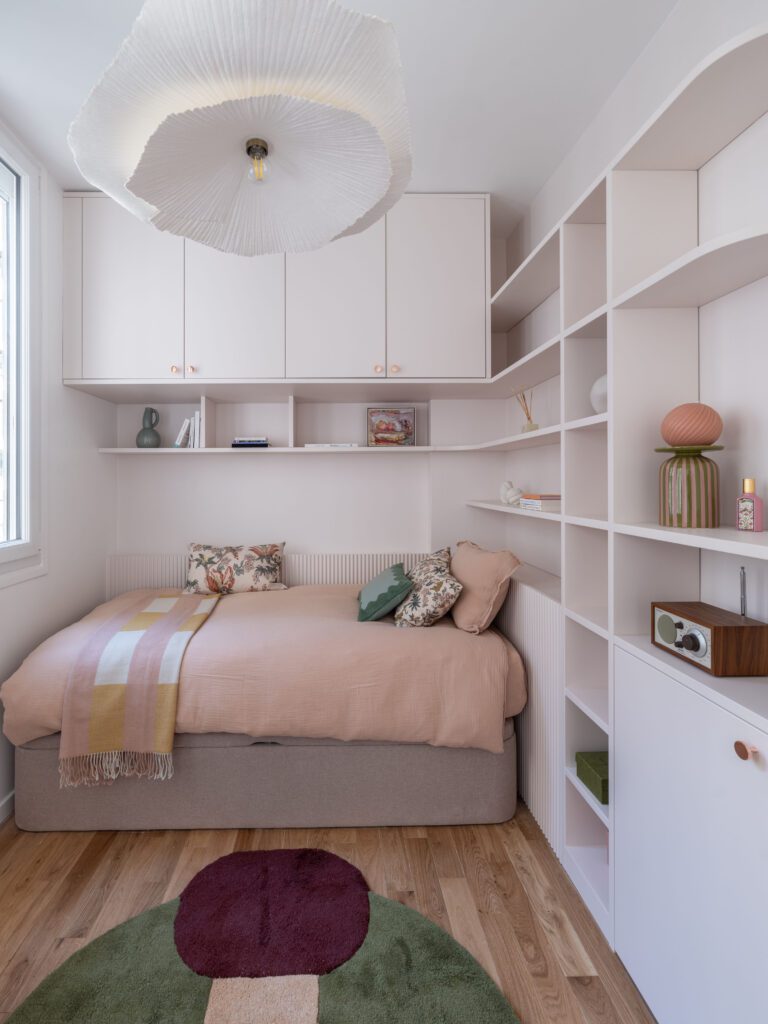 Chambre d'adolescente aménagée avec soin dans un appartement parisien rénové, mettant en valeur un lit confortable avec couverture rose poudré et coussins décoratifs. Rangements sur mesure couleur rose arum avec étagères intégrées, miroir organique et luminaire suspendu élégant. Décoration intérieure moderne et fonctionnelle, alliant couleurs douces, rangements sur mesure optimisés et accessoires décoratifs pour un espace à la fois esthétique et ergonomique.