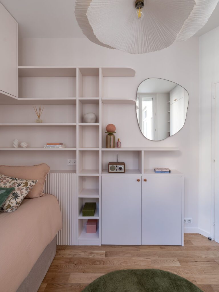 Chambre d'adolescente aménagée avec soin dans un appartement parisien rénové, mettant en valeur un lit confortable avec couverture rose poudré et coussins décoratifs. Rangements sur mesure couleur rose arum avec étagères intégrées, miroir organique et luminaire suspendu élégant. Décoration intérieure moderne et fonctionnelle, alliant couleurs douces, rangements sur mesure optimisés et accessoires décoratifs pour un espace à la fois esthétique et ergonomique.